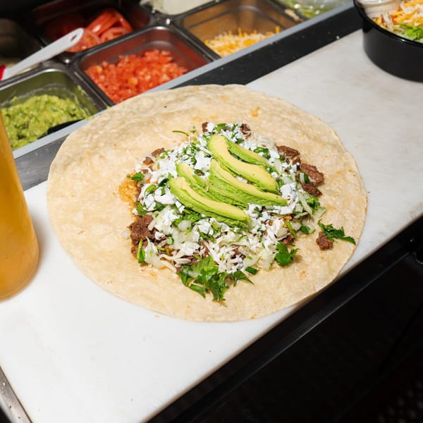 A large El Flamin Taco burrito being assembled with meat, avocado, and other toppings.