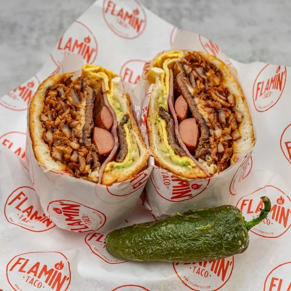 A large El Flamin Taco torta, cut in half, revealing meat, hot dogs, avocado, and egg, with a jalapeño.