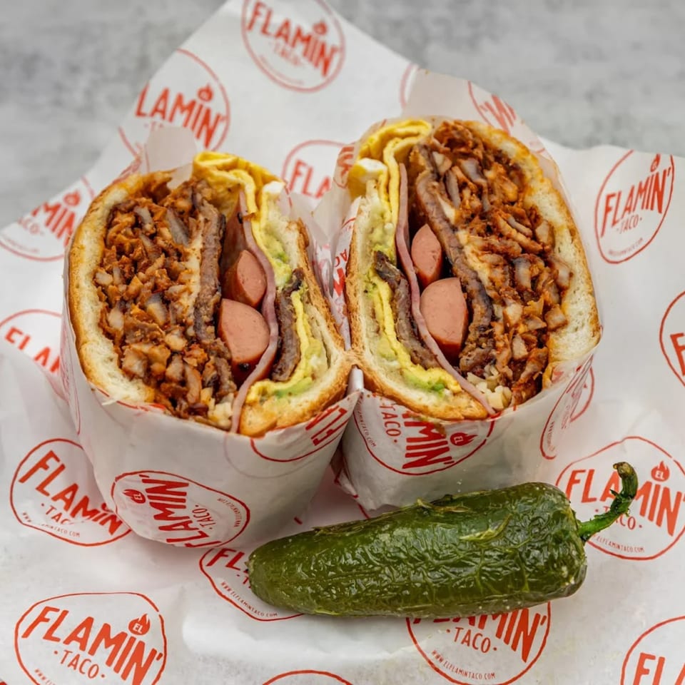 A large El Flamin Taco torta, cut in half, revealing meat, hot dogs, avocado, and egg, with a jalapeño.