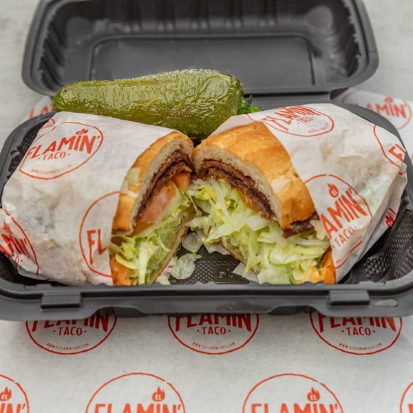 A sliced El Flamin Taco sandwich with meat, lettuce, and tomato, alongside a pickle in a takeout container.