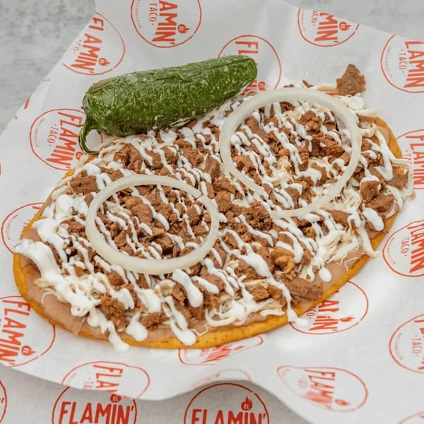A large El Flamin Taco huarache with meat, crema, onions, and a whole jalapeño on branded paper.