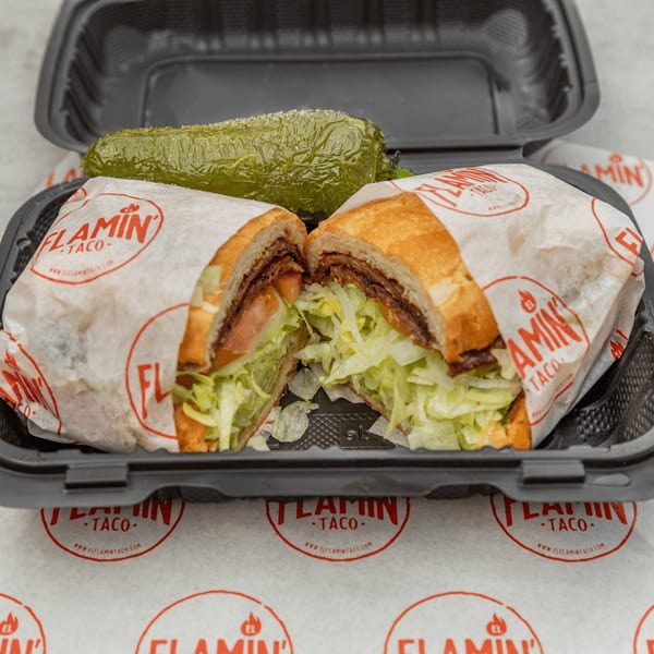 A sliced El Flamin Taco sandwich with lettuce and tomato, next to a pickle in a black to-go container.