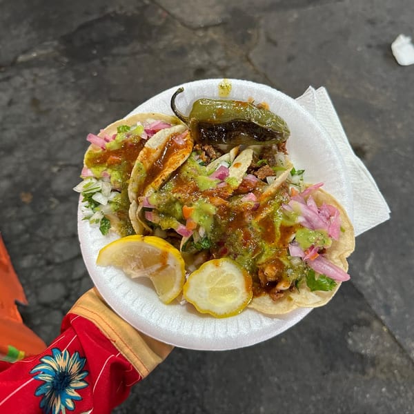 A hand holds a white plate with four El Flamin Taco street tacos, lemon slices, and a grilled pepper.