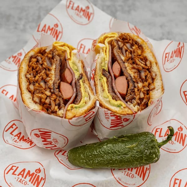 A large El Flamin Taco torta, cut in half, revealing various meats, avocado, and hot dogs.